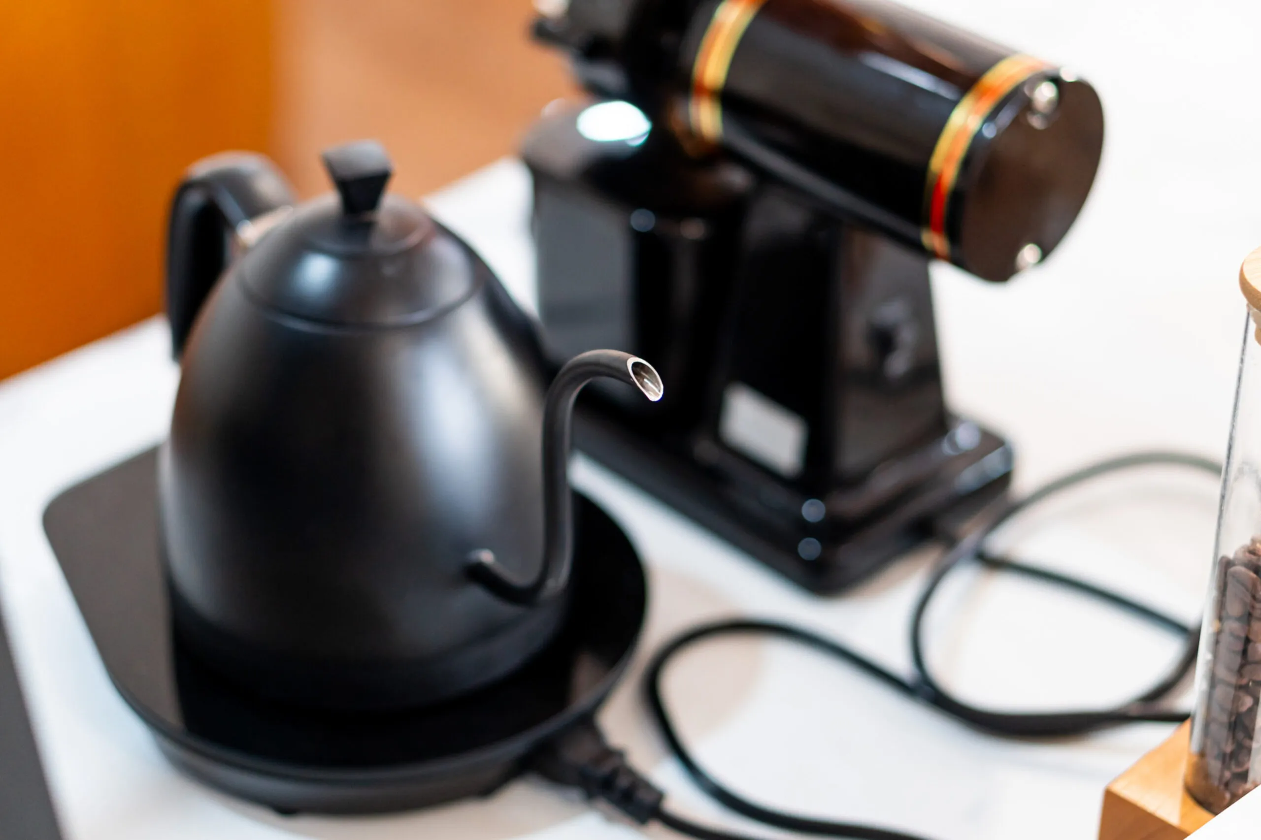 A sleek black kettle with a long spout sits on a black base, next to a coffee grinder. The kettle has a modern design, and the grinder features a shiny black finish with